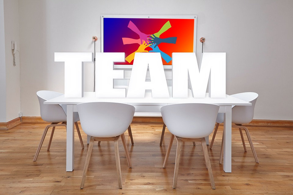 Office conference table with large letters spelling out “TEAM”. Behind the letters is a colourful painting on the wall with 5 hands overlapping together.
